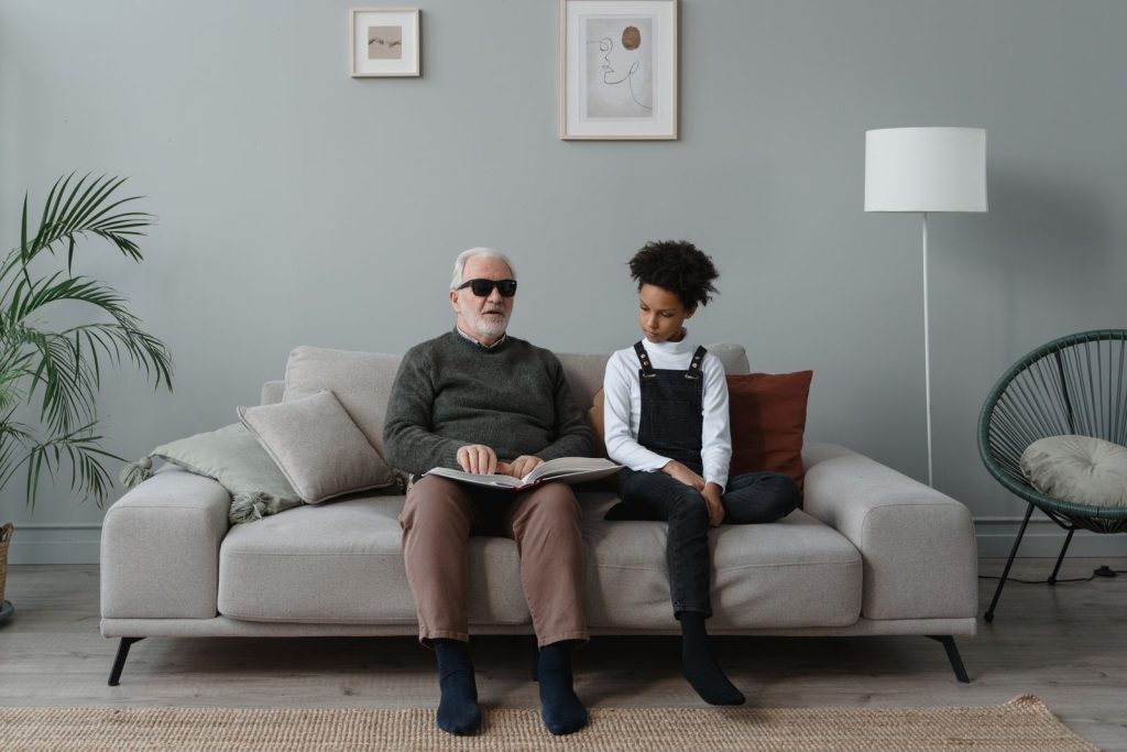 A senior man with sunglasses reads Braille with a young boy, seated on a cozy sofa.