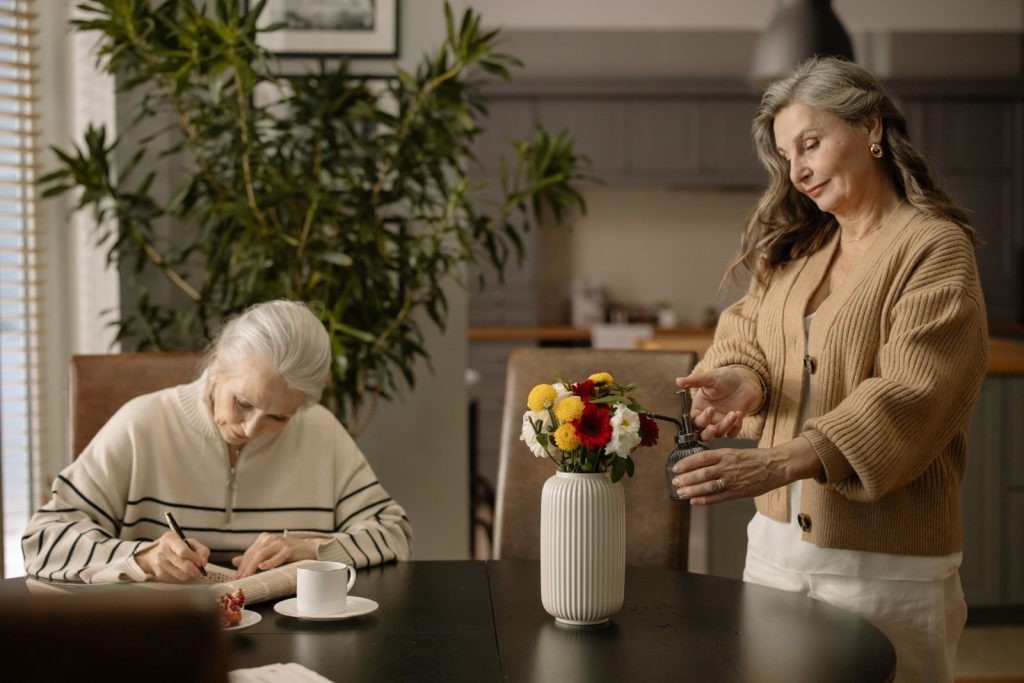 Two senior women enjoy a cozy afternoon in a dining room with a flower arrangement.