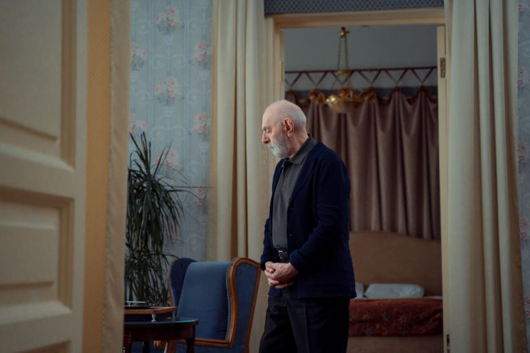 Elderly man standing thoughtfully in a vintage styled room with elegant decor.