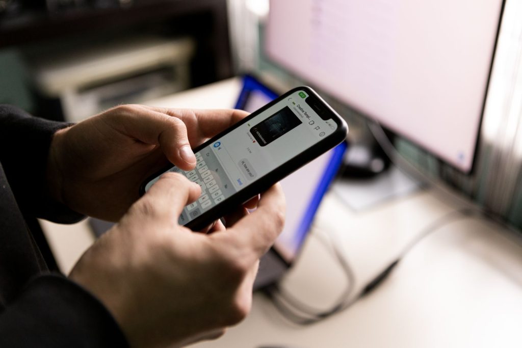 A person holding a smartphone and chatting indoors near a computer screen.
