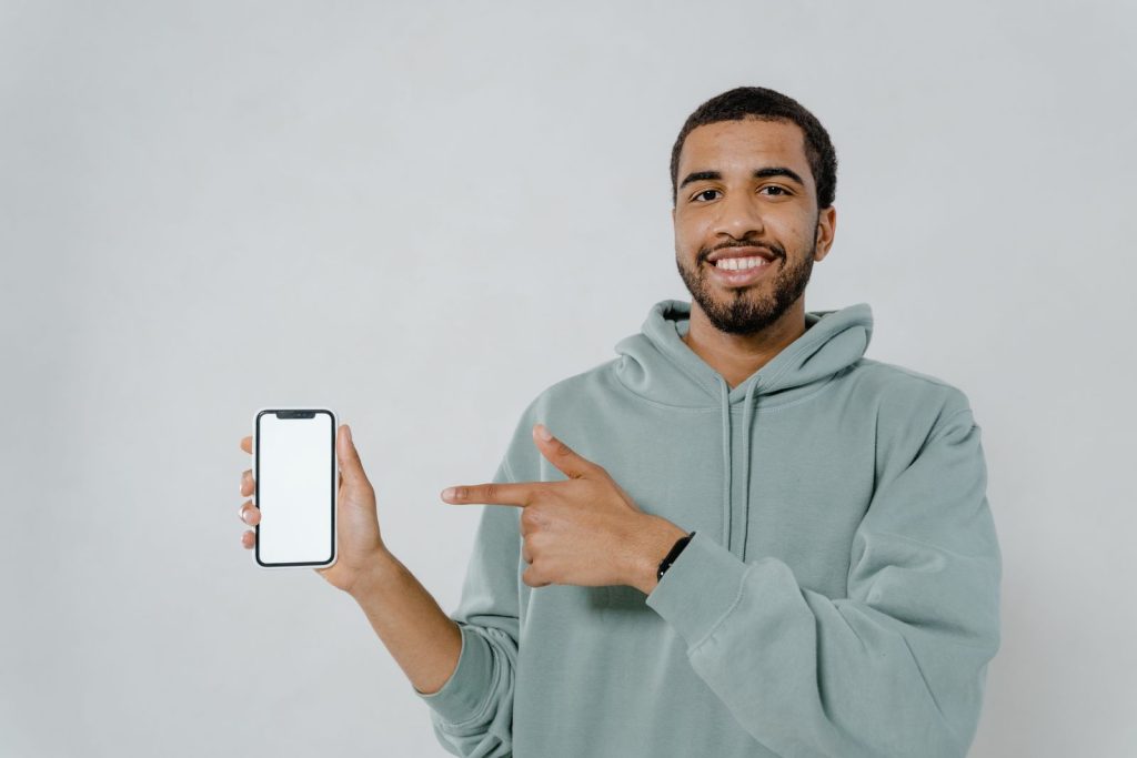 A smiling man in a hoodie points at a smartphone with a blank screen, ideal for mockup or design use.