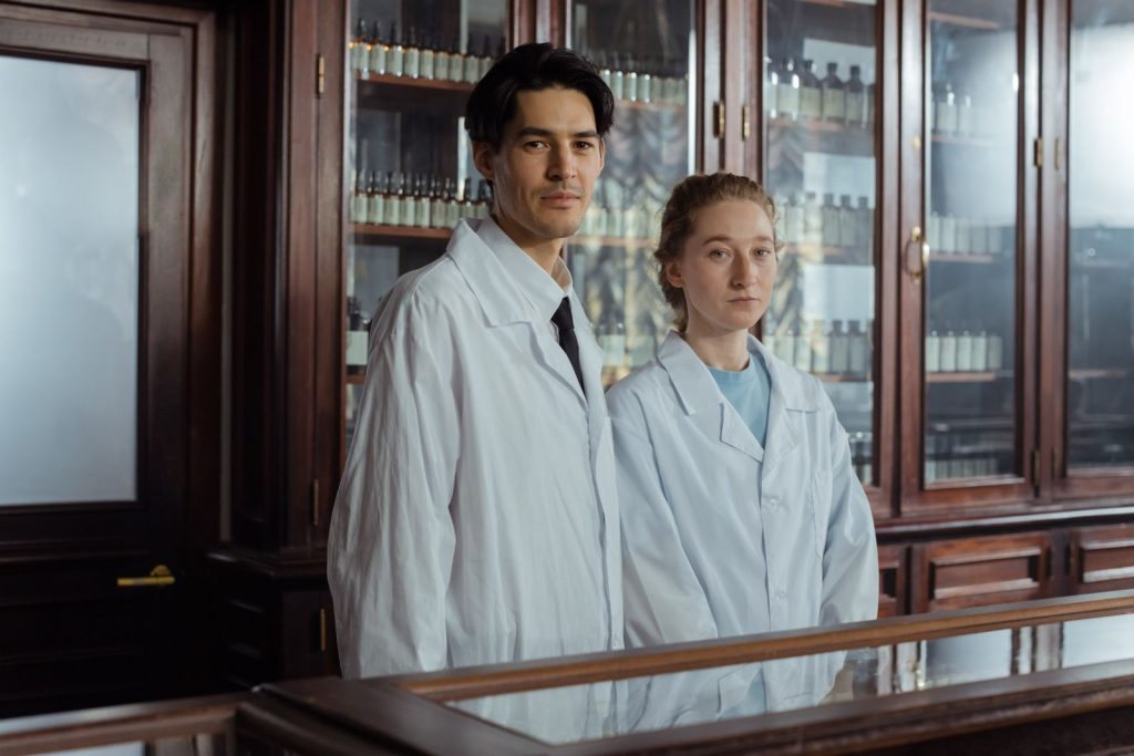 Two pharmacists in white gowns stand in an antique pharmacy with vintage cabinets.