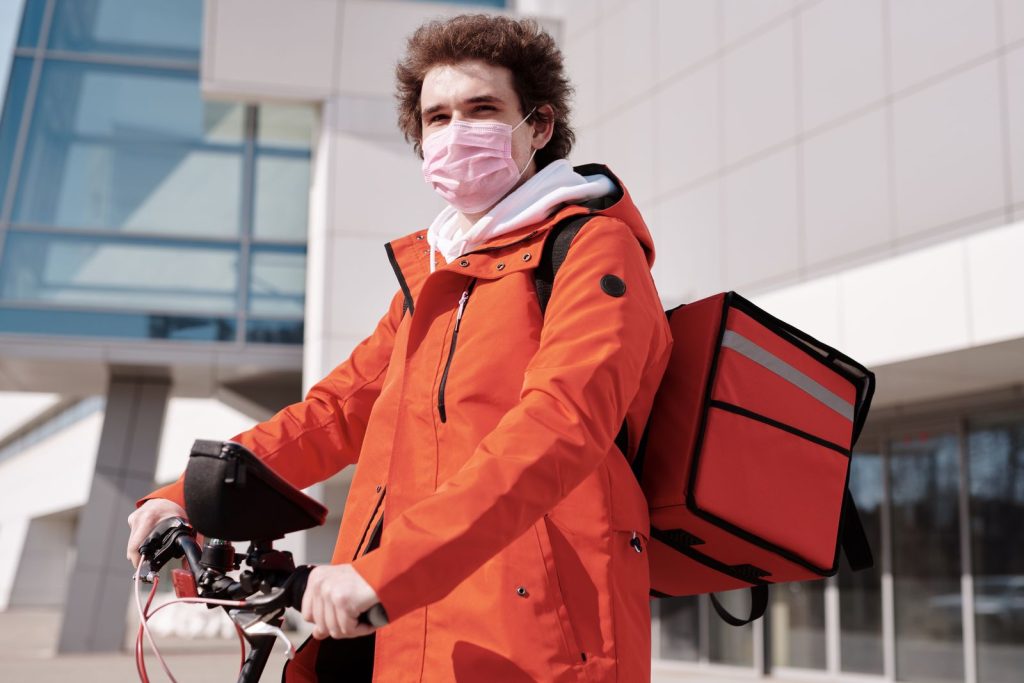 A delivery person on a bike wearing a mask and carrying a thermal bag, outdoors.