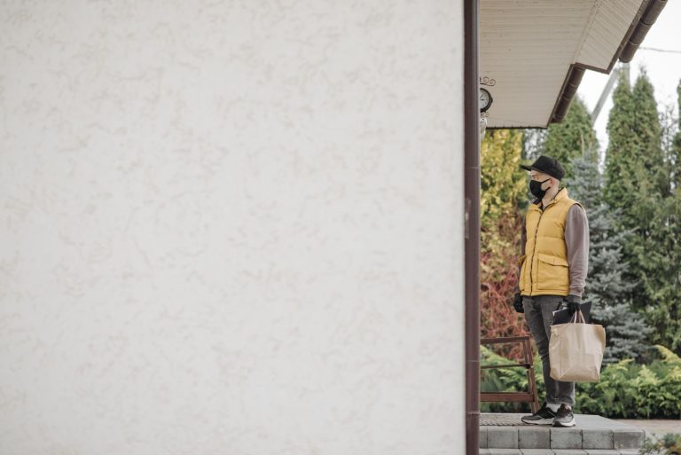 Delivery man wearing a facemask and carrying a paper bag, standing outside a house.