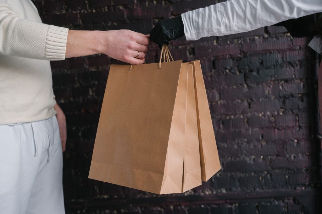 Two individuals exchange paper shopping bags against a dark brick wall background.