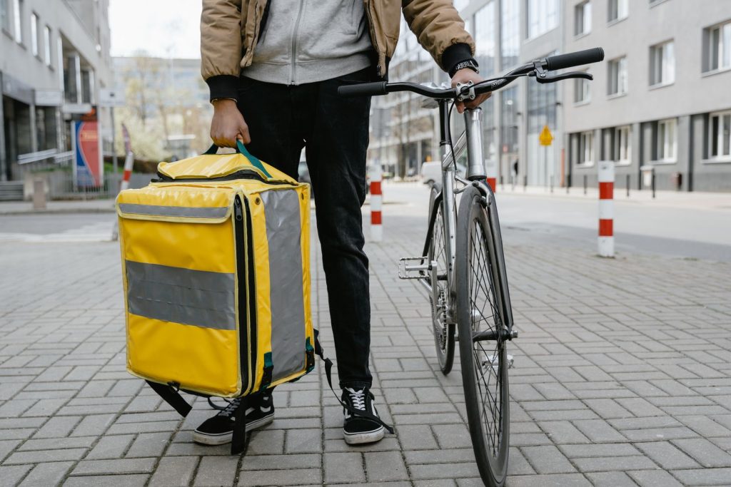 A cyclist with a yellow thermal bag stands with his bike on a city street, ready for food delivery.