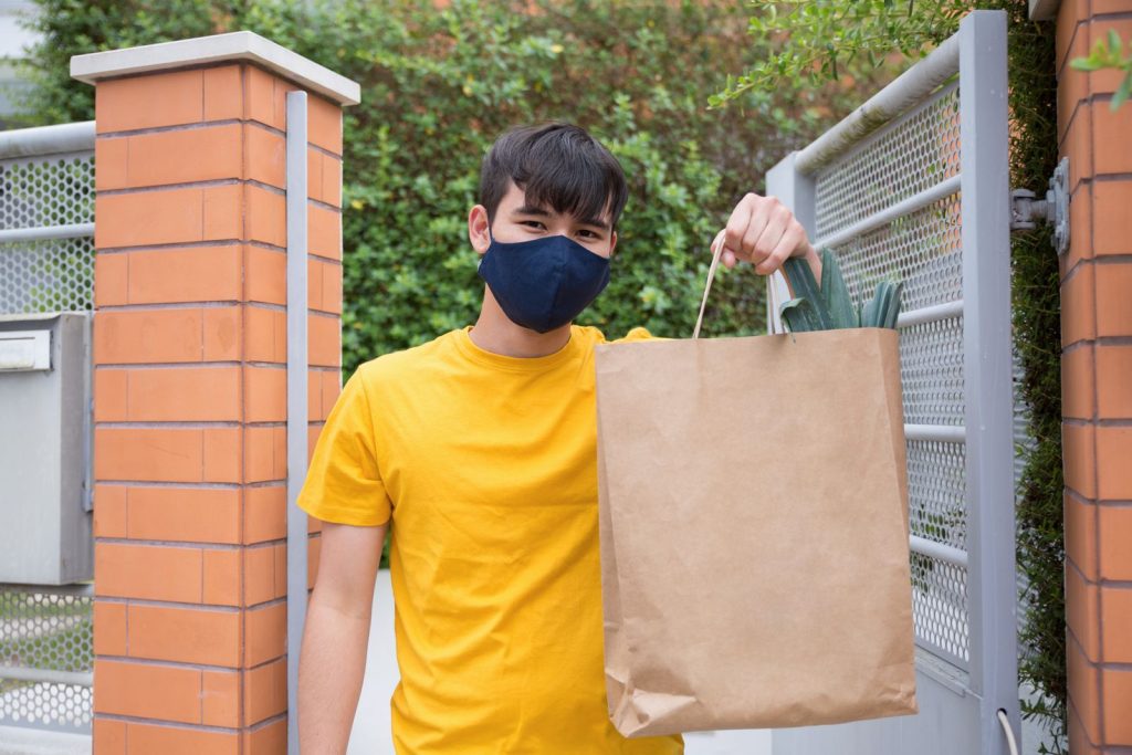 A young man wearing a mask holds a paper bag with groceries outside a gate on a sunny day in Portugal.