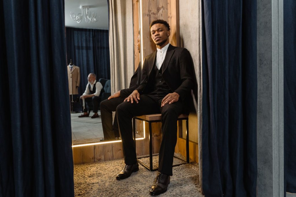 A stylish man in a suit sits in a tailor shop fitting room, embodying elegance and sophistication.