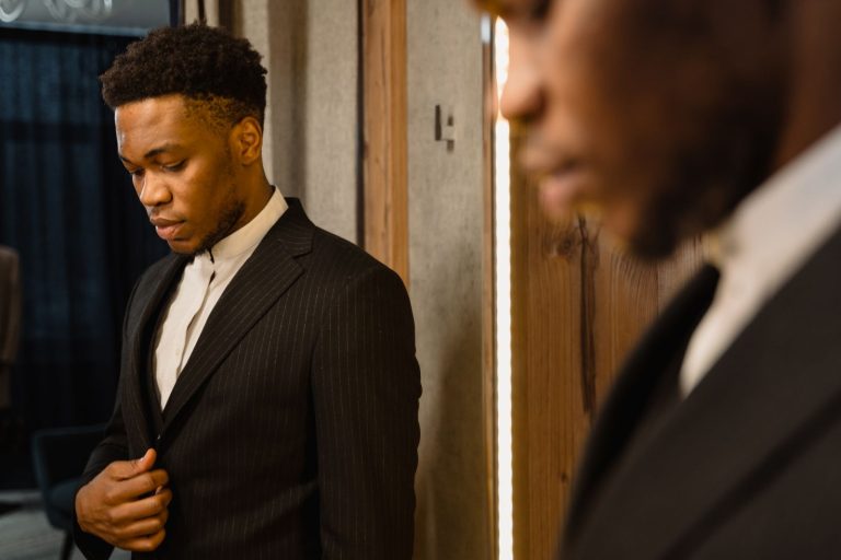 Stylish man in a suit ponders in front of a mirror indoors, captured in warm lighting.