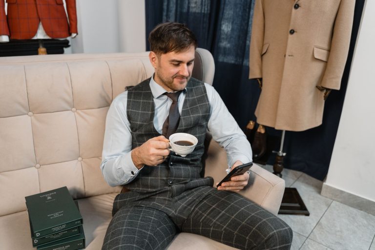 Man in a plaid suit enjoying coffee and checking his mobile phone while relaxing on a sofa.