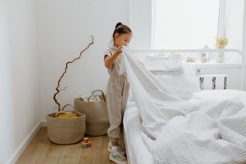 A young girl making her bed in a bright, minimalist bedroom.