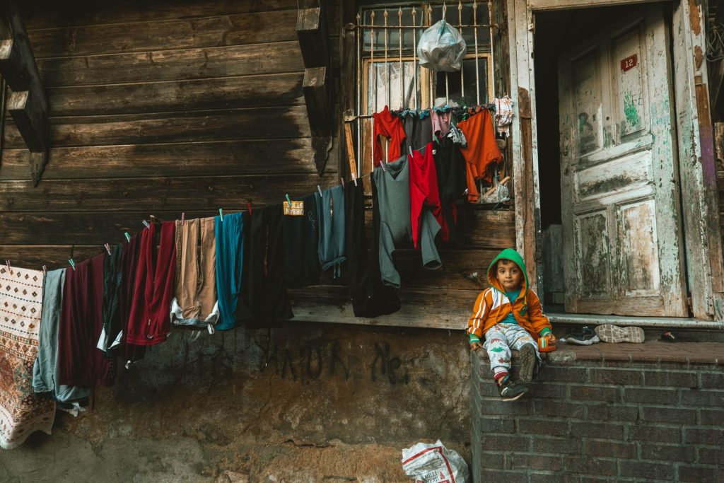 A child in a colorful hoodie sits outside a rustic wooden house in Istanbul. Clothes hang to dry.