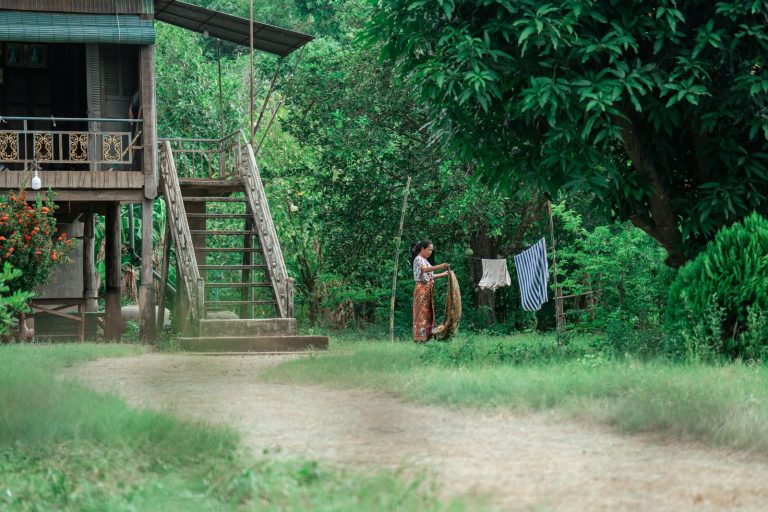 A woman drying clothes near a traditional wooden house surrounded by lush greenery in a rural setting.