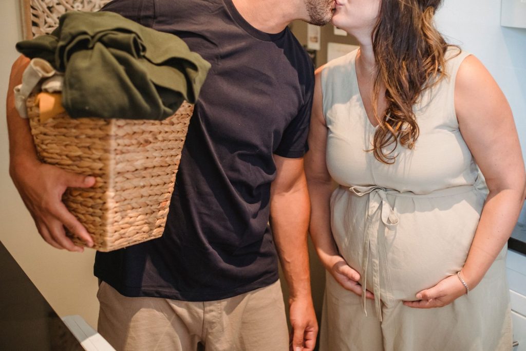 Unrecognizable adult pregnant woman kissing man with laundry basket while standing together in room