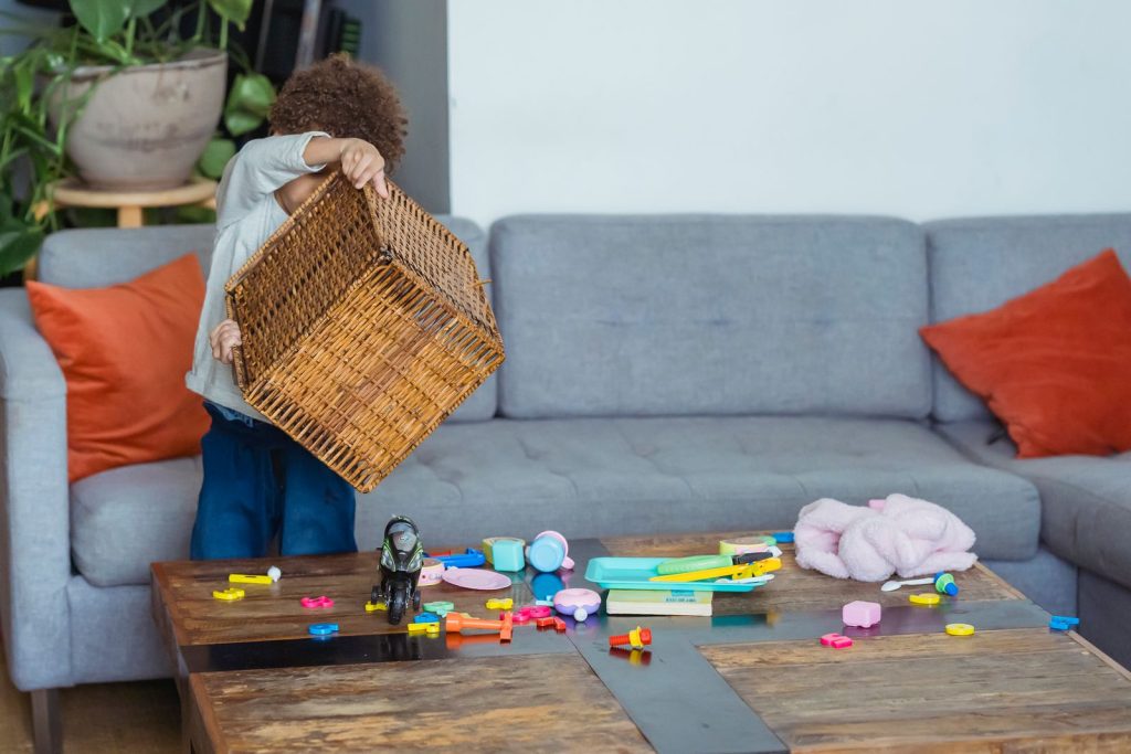 Ethnic kid in jeans standing near sofa and table and playing with colorful details from overturned braided box