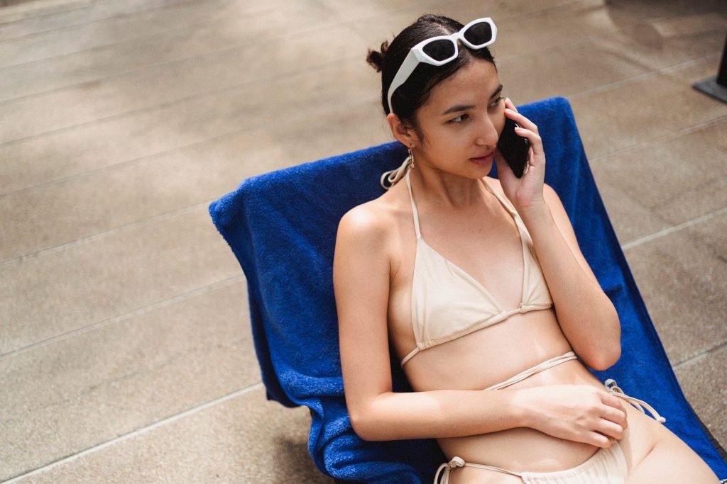 From above of young relaxed ethnic lady talking on mobile phone while lying on sunbed in stylish bikini and sunglasses during summer holidays