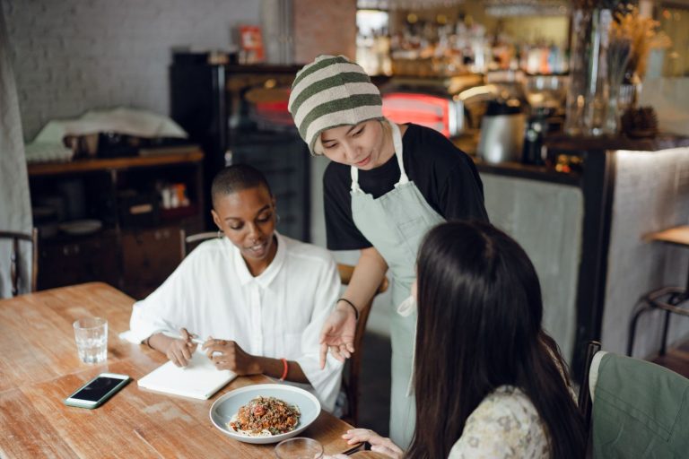 Positive smiling multiethnic colleagues in formal wear making order to ethnic waitress in apron while sitting at wooden table in contemporary cafe in daytime