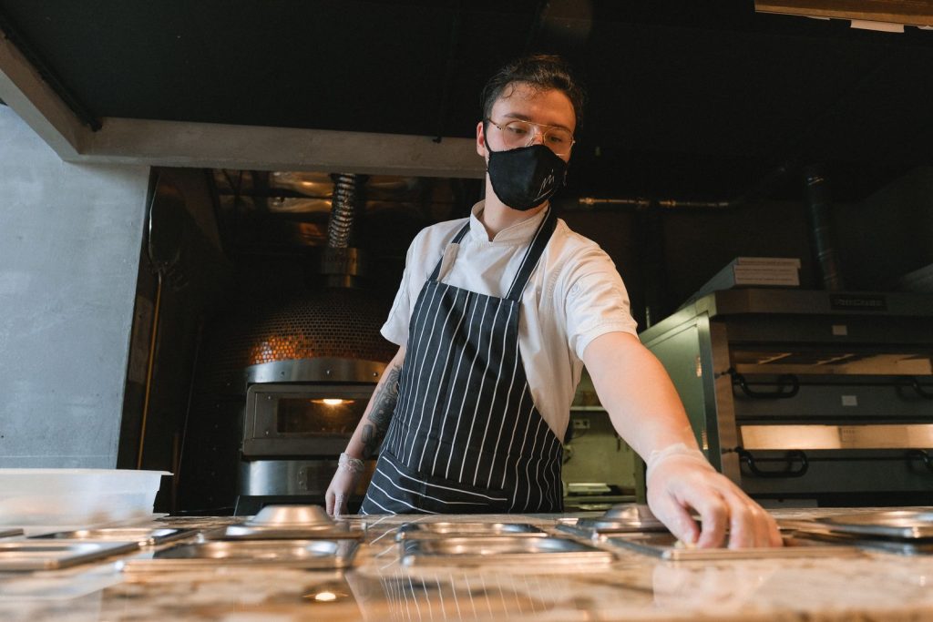 A baker wearing a face mask and apron works in an industrial kitchen setting.