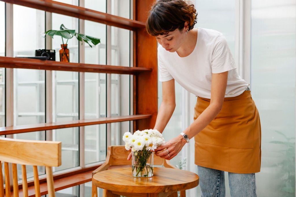 Crop young female cafeteria worker putting vase with blossoming flowers on wooden table in daytime