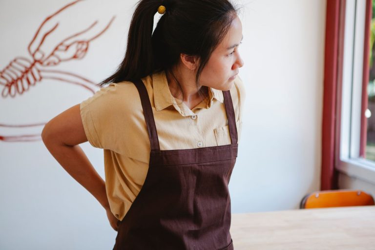 Asian woman in a brown apron standing near a window with a neutral background.