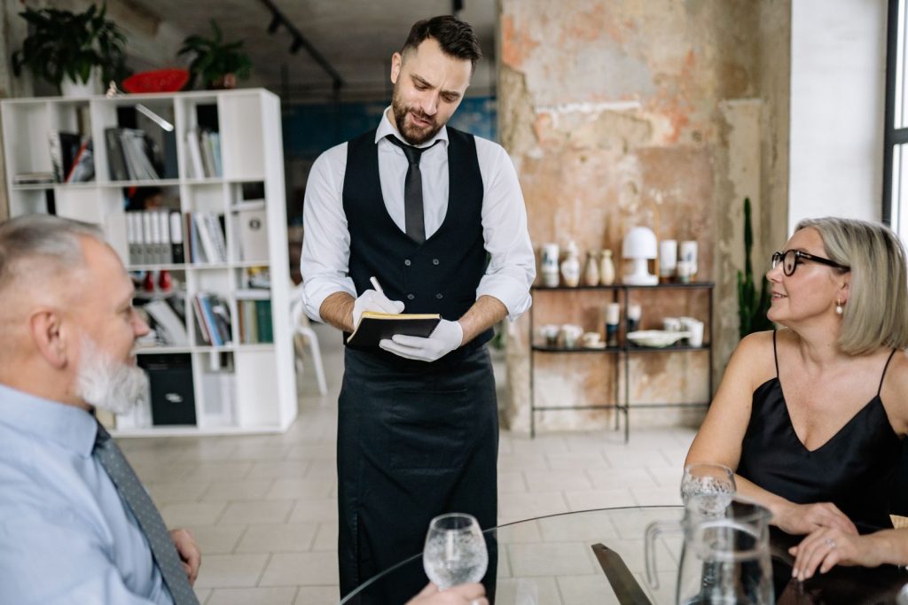 A waiter takes orders from a senior couple in a stylish, modern restaurant interior.