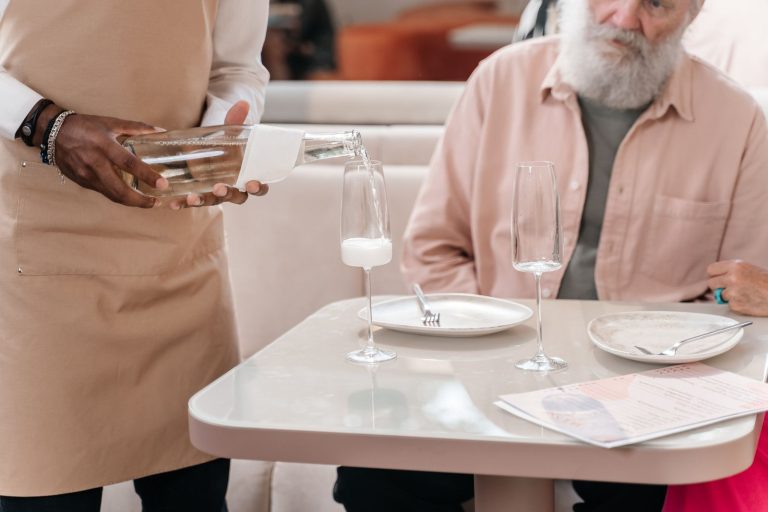 A waiter pouring a drink for an elderly man seated at a restaurant table. Focus on hospitality.