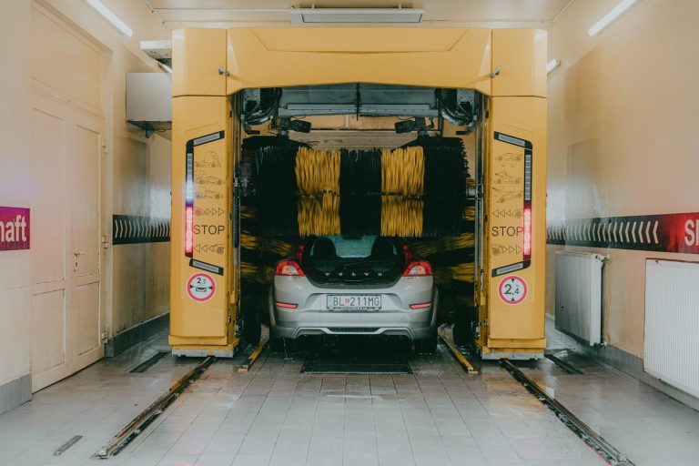 A car undergoing a cleaning process in an automated car wash station with a yellow gantry machine.