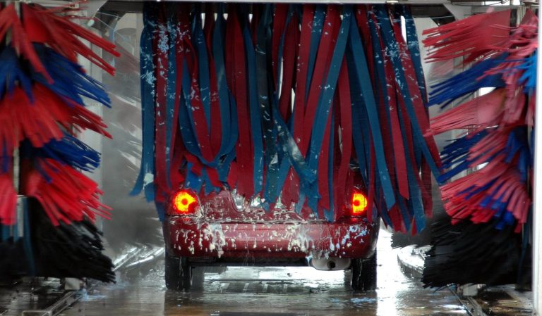 Red car being cleaned in an automated car wash with colorful brushes and soap suds.