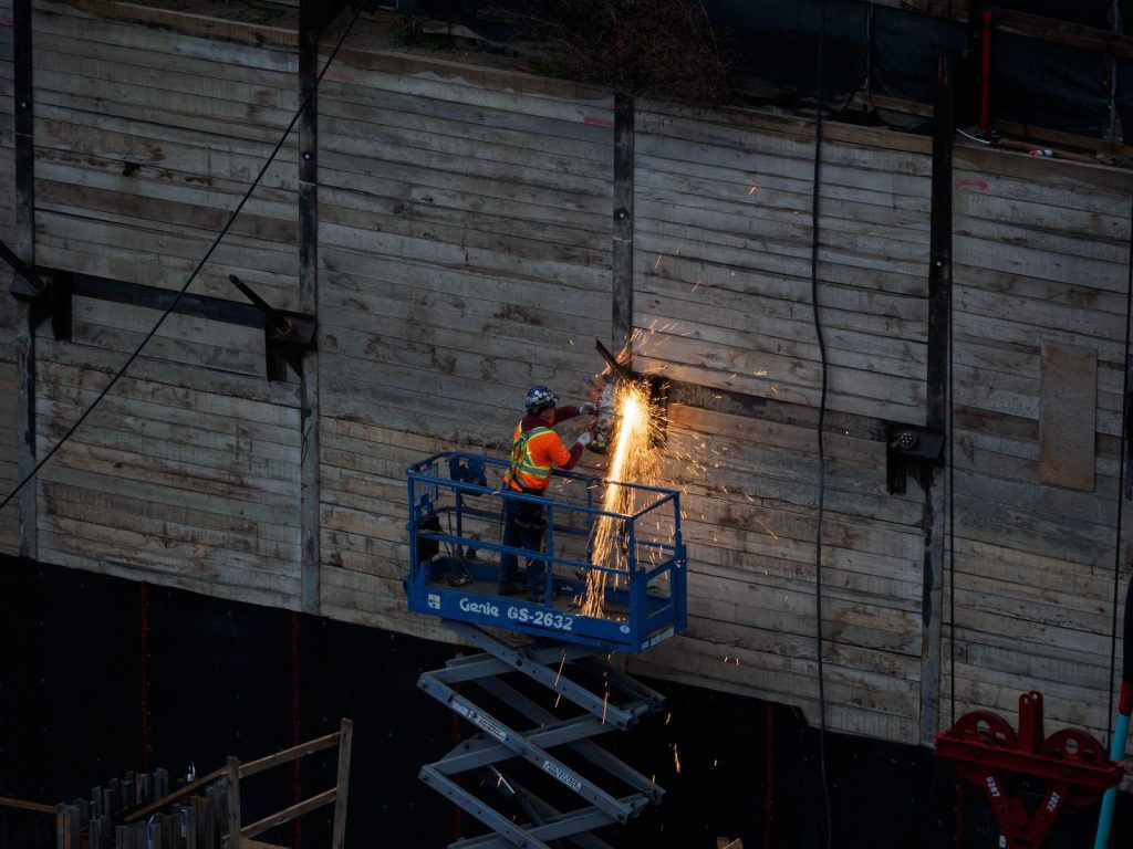 A construction worker welds on a raised platform, creating sparks on an industrial site.