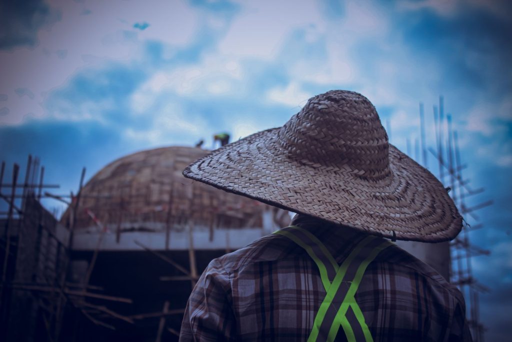 A construction worker wearing a straw hat observes a construction site under a blue sky.