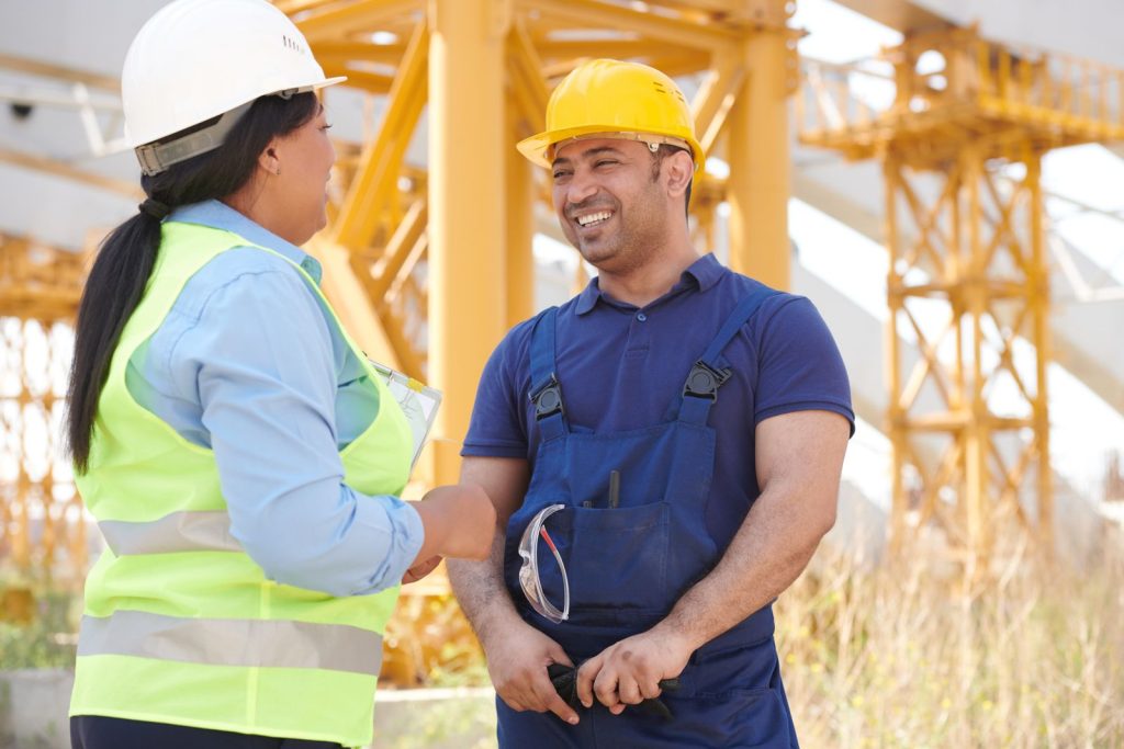 Two construction workers in hardhats smiling and talking on a sunny day at a site.