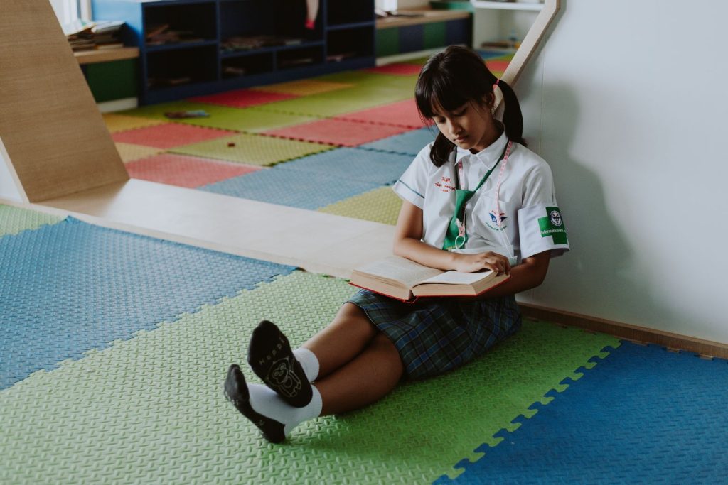 A young girl in school uniform sits on colorful mats reading a book indoors.