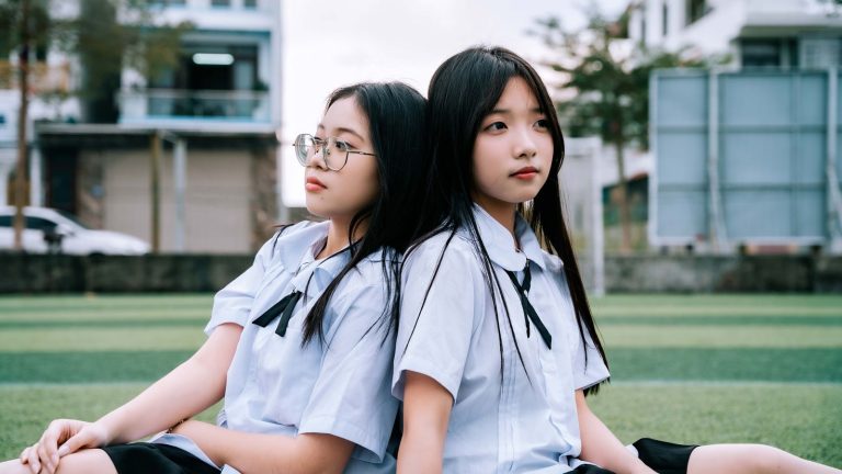 Two schoolgirls in uniforms sitting on a field, school background, thoughtful expressions.
