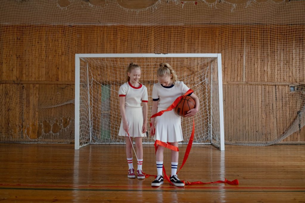 Two young girls standing in a gym celebrating with a basketball and ribbon.