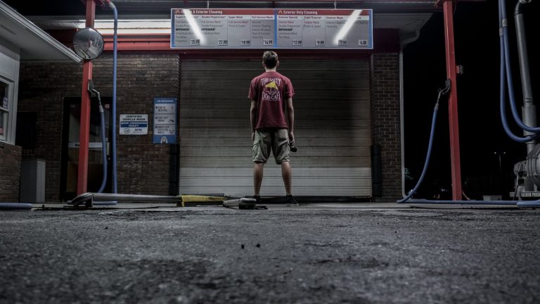 A man stands at a self-service car wash at night, illuminated by overhead lights.