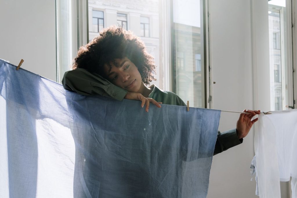 A thoughtful woman in natural light leaning on a clothesline indoors with calm expressions.