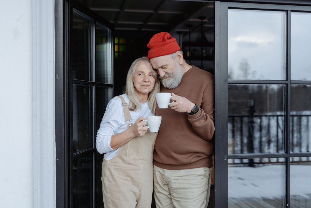 Happy senior couple stands with coffee by the door on a snowy winter day.