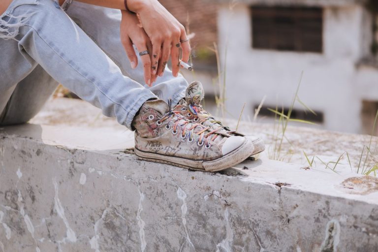 Teenager in distressed jeans sitting outdoors, wearing vintage Converse sneakers and holding a cigarette.