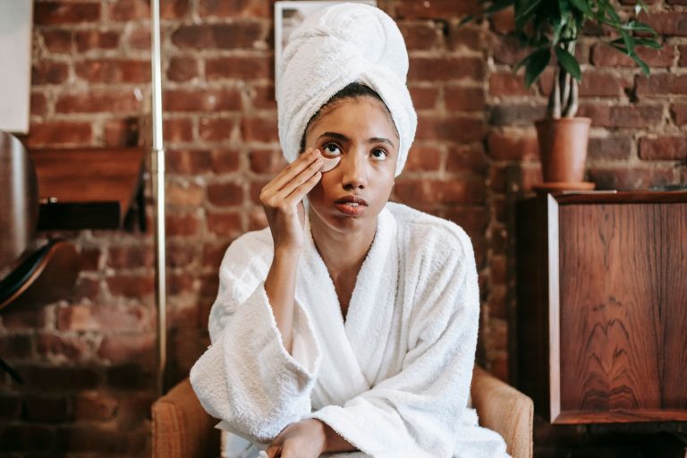 Young female in white bathrobe and towel turban applying moisturizing facial patches while sitting on armchair in kitchen