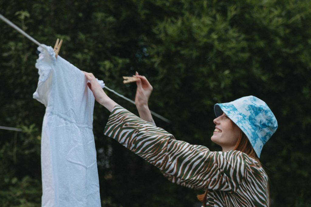 Smiling woman hanging white dress on clothesline in garden, enjoying a sunny day.