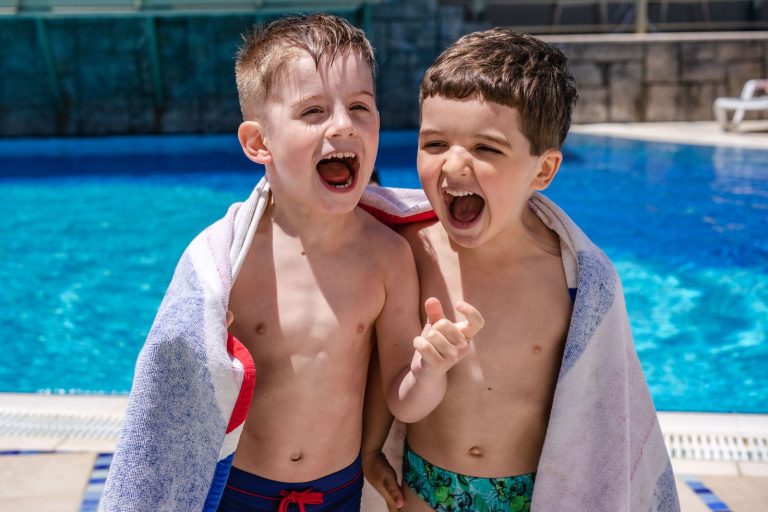Two happy caucasian boys laughing by the pool, wrapped in towels during a sunny day.