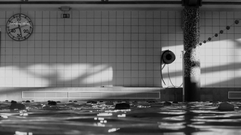 Monochrome view of indoor swimming pool with clock and tiles.