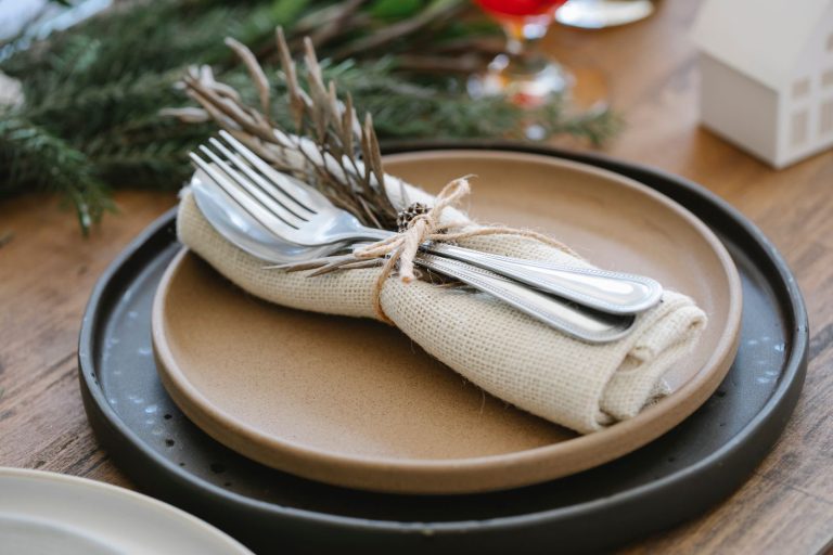 From above of plates with cutlery and napkin tied by ribbon with dry twig placed on wooden table near coniferous branch