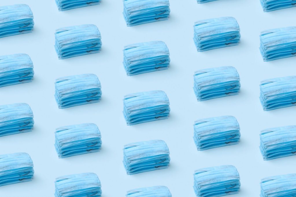 Stacks of blue medical face masks arranged in a repeating pattern on a light blue background.
