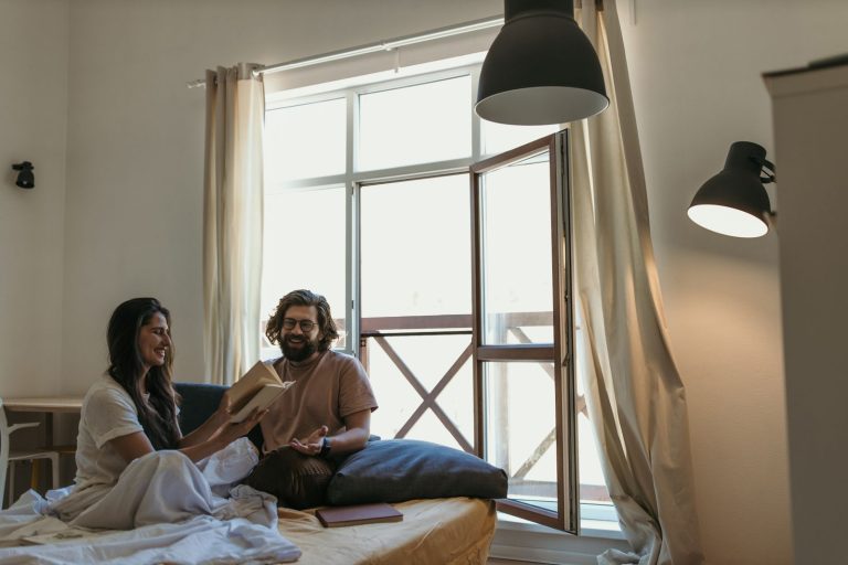 A cheerful couple laughing and sharing a book in a cozy bedroom setting.