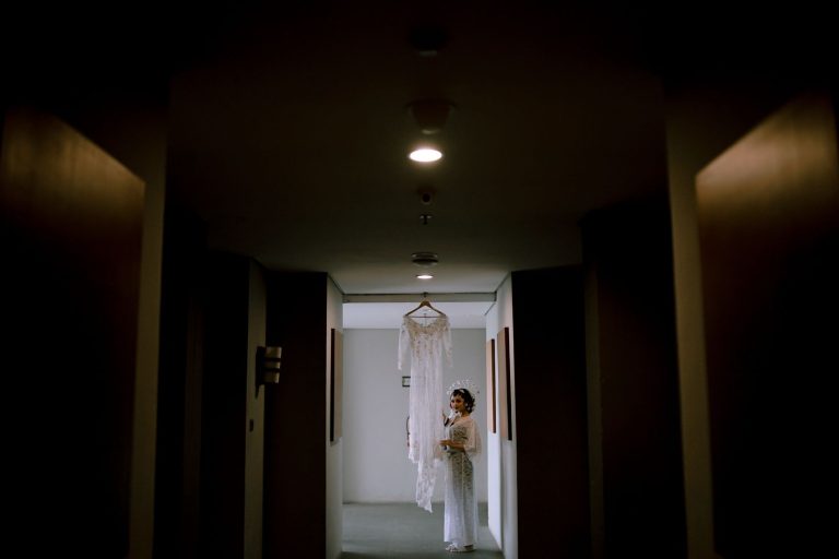 A bride stands in a dark corridor, admiring her wedding dress hanging in the light.
