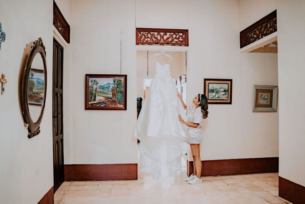A woman admires her elegant wedding dress hanging in a classic, art-adorned room.