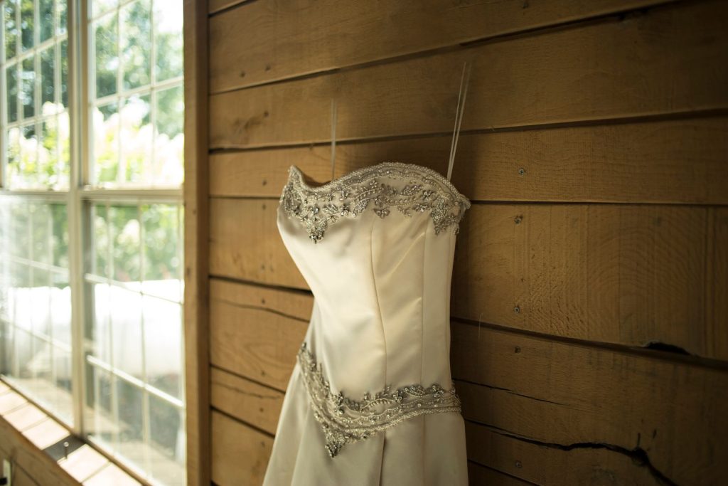 A beautifully embroidered wedding dress hanging on a rustic wooden wall by a window, evoking elegance and charm.