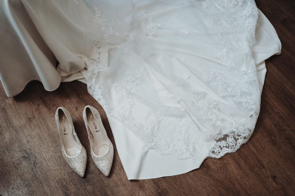 Close-up of a white bridal gown with lace and sparkling shoes on wooden floor.