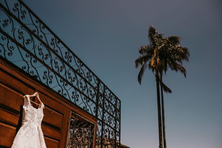 A beautiful white wedding gown hangs outdoors beside a palm tree and ornate fence.